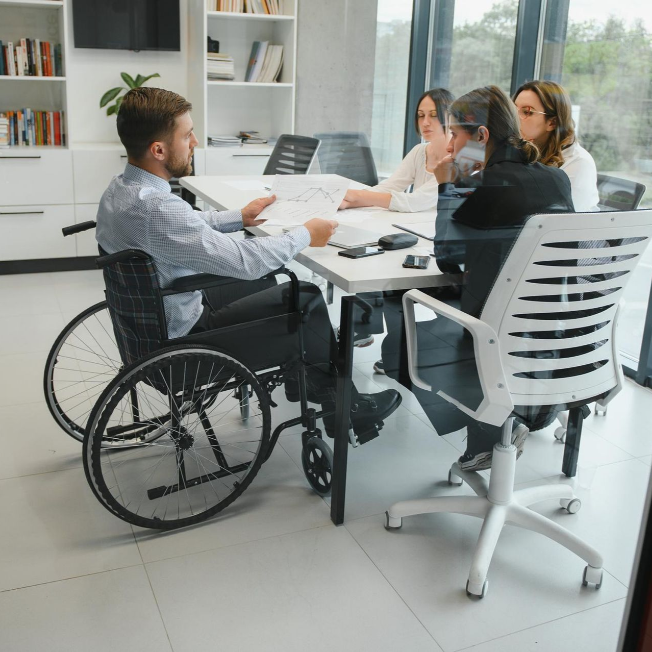 Wheelchair user in a bright office meeting with three colleagues around a conference table.