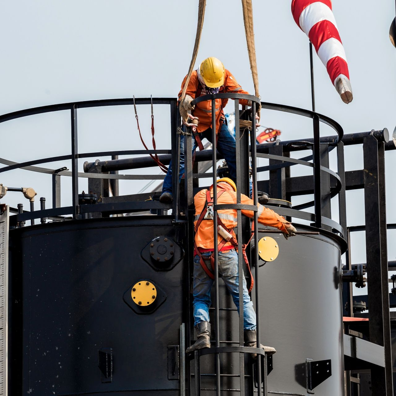 Two workers in orange safety gear climb a ladder on an industrial tank with a crane overhead