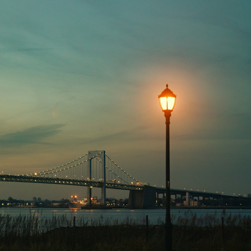 Lit streetlamp by a suspension bridge at dusk over calm water