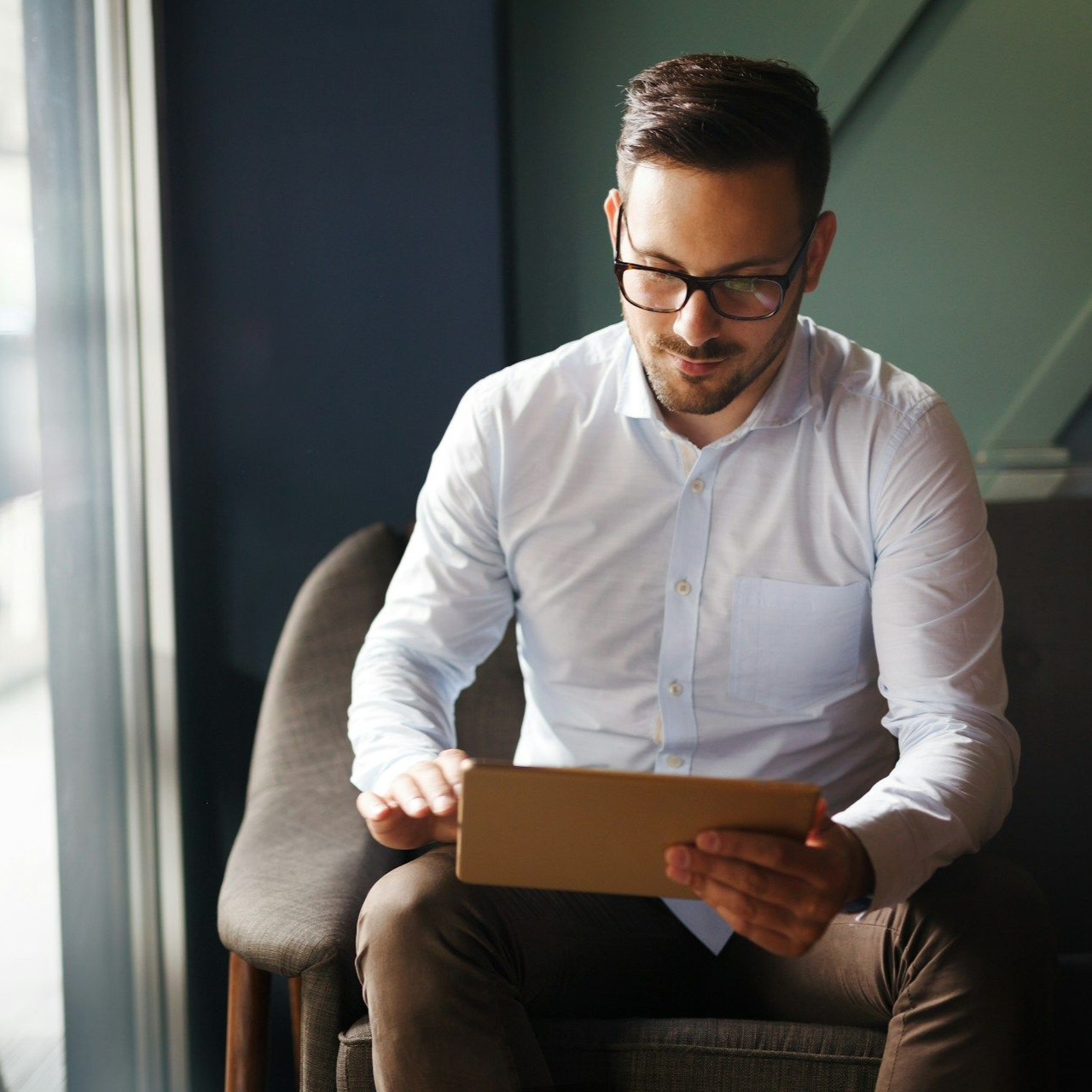 Man in glasses seated by a window, looking at a tablet in a bright office.