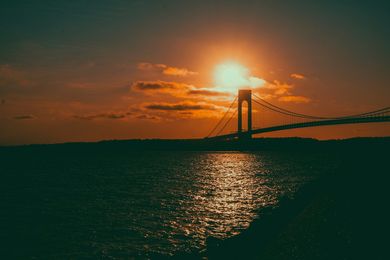 Sunset over a suspension bridge, with orange sky reflected on calm dark water.