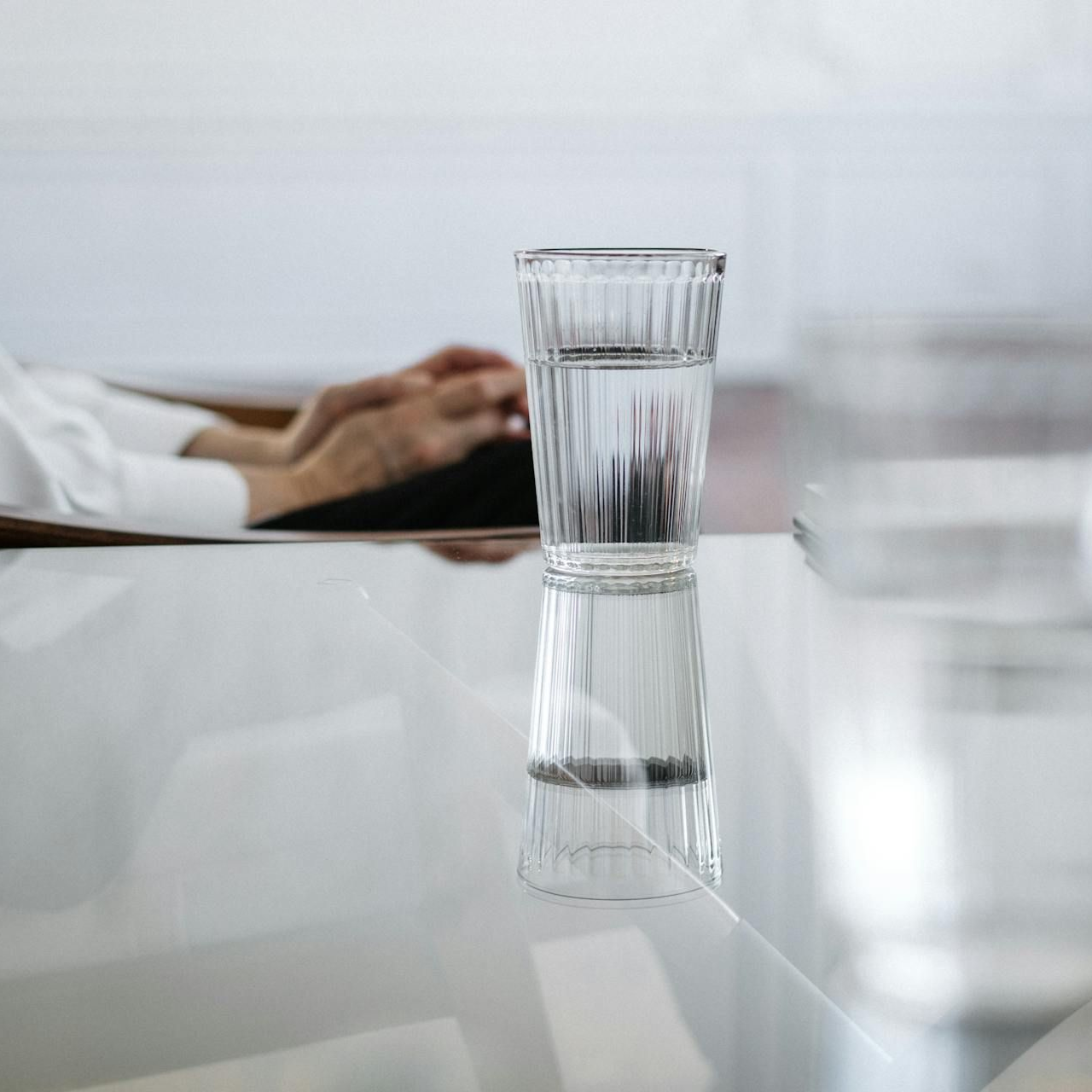 A glass of water on a reflective table with a blurred hand in the background