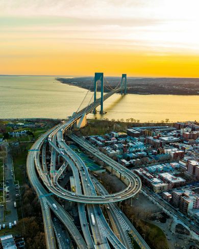 Suspension bridge and highway interchange at sunset over a wide river, with city buildings along the shore