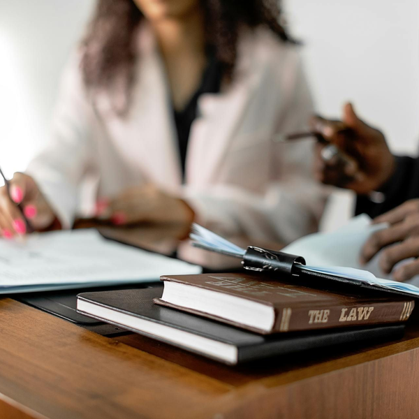 Stacked notebooks and a pen on a wooden table during a meeting, with people blurred in the background