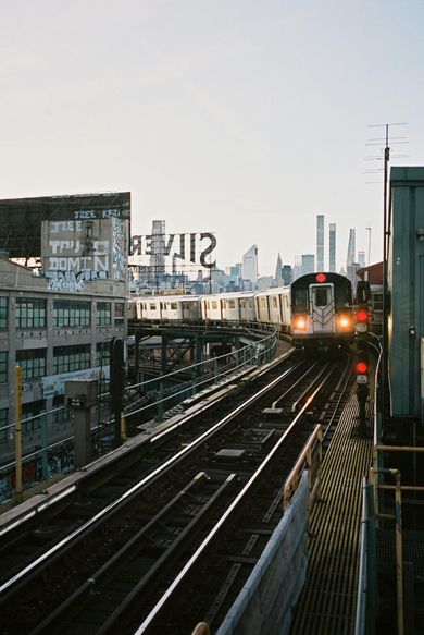 Subway train approaching elevated tracks in a city at dusk, with buildings and graffiti beside the line