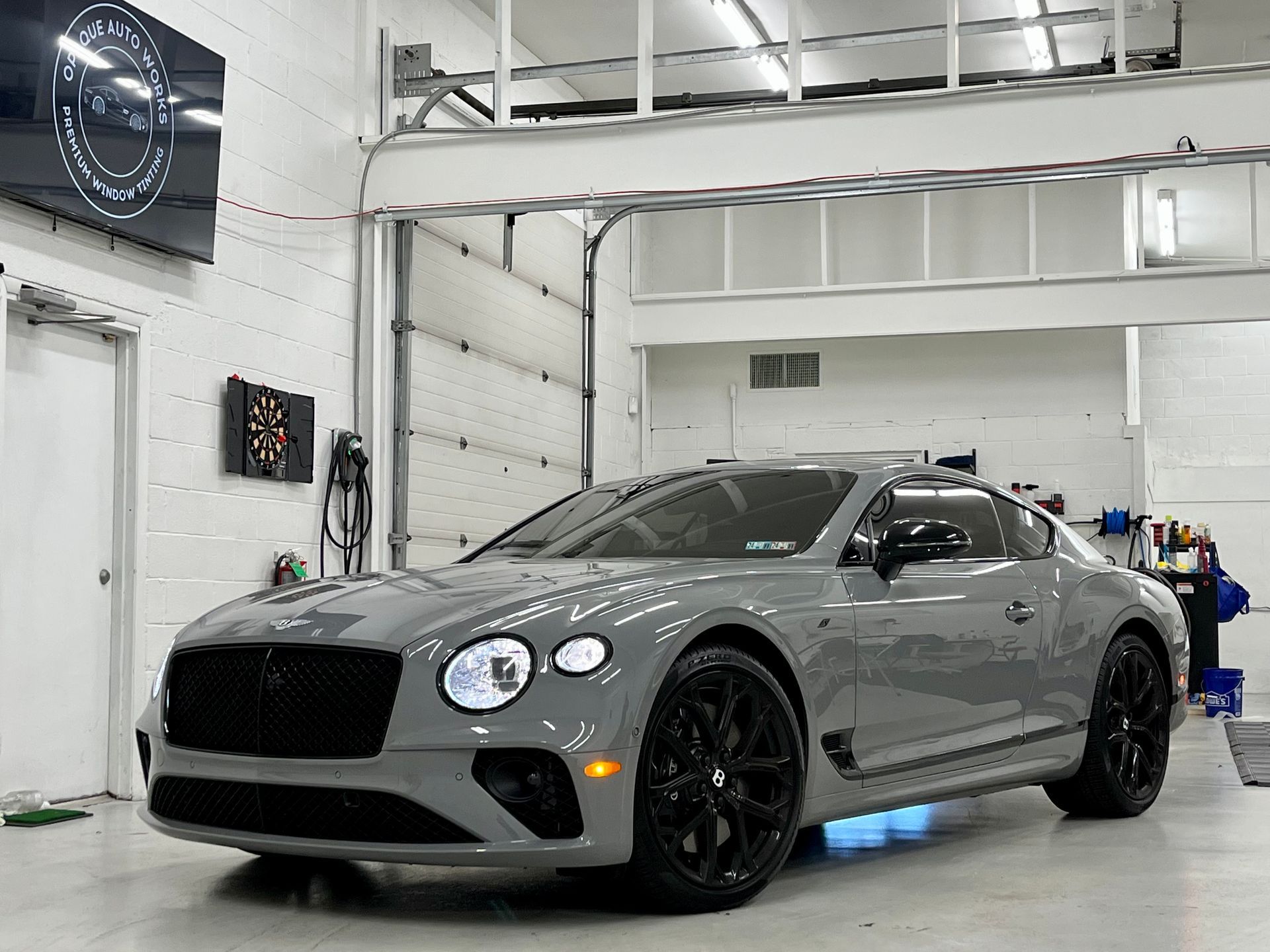 Gray Bentley coupe parked inside a brightly lit garage. Black wheels, headlights on.