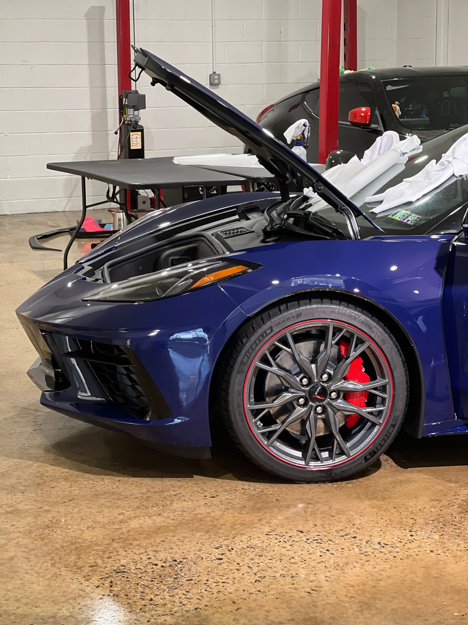 Blue sports car with hood open in a garage, red brake calipers visible on the front wheel.