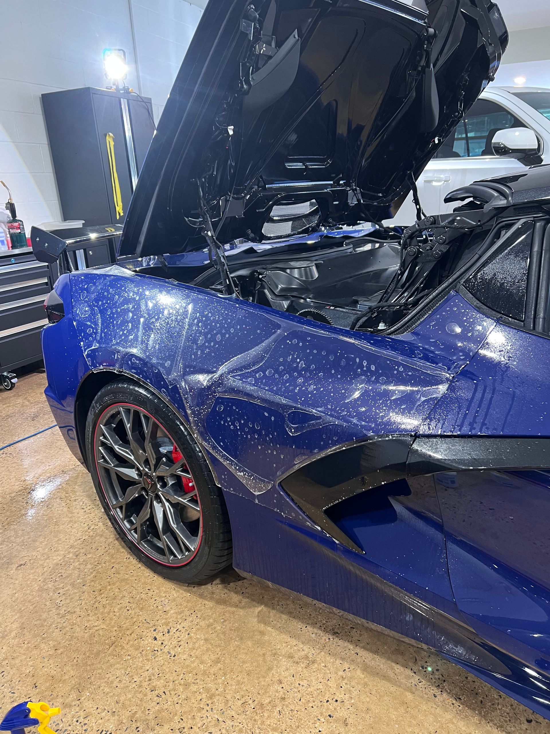 Blue sports car being washed; hood up, water droplets visible. Black wheels with red accents.
