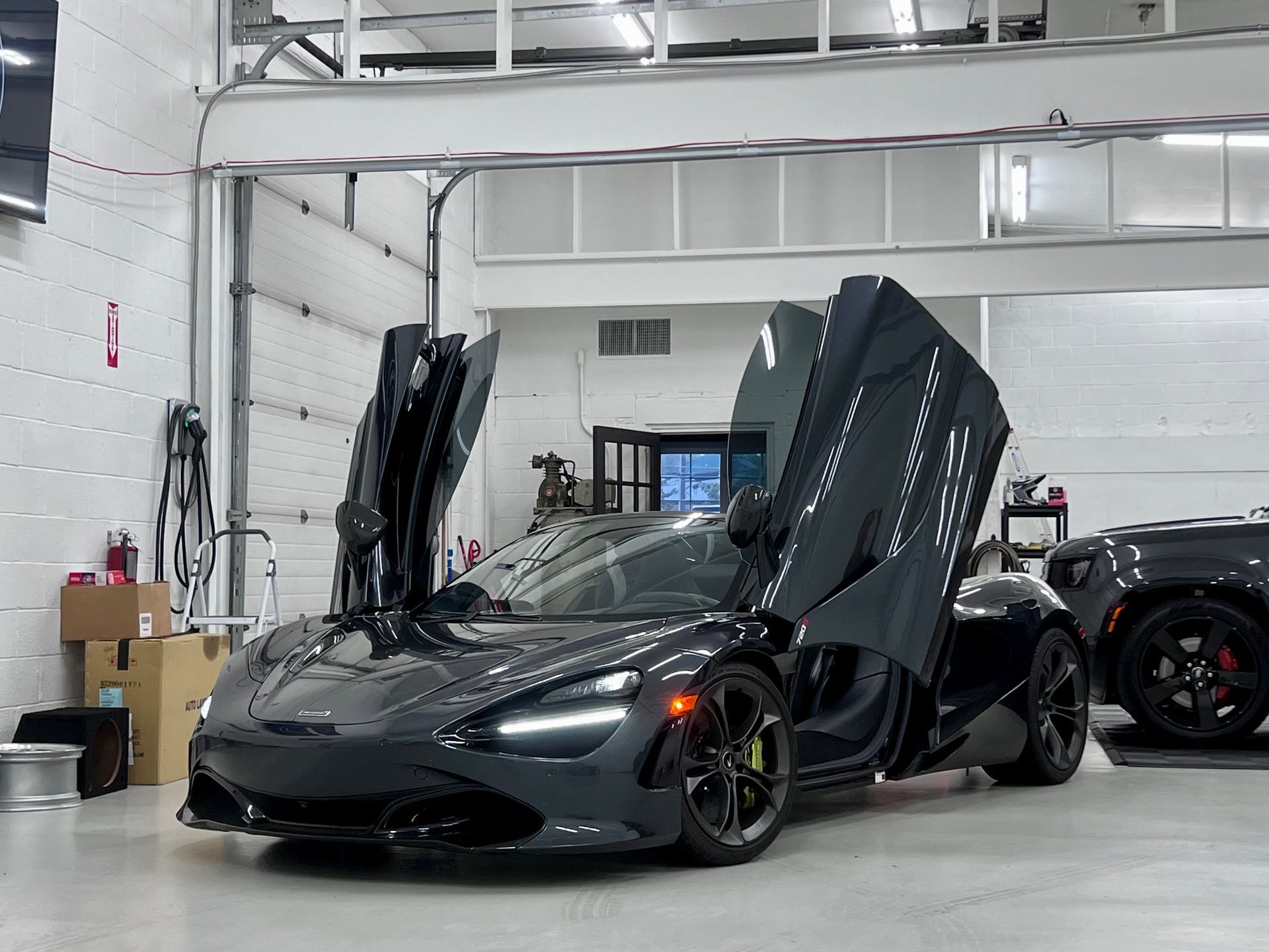 Black sports car with butterfly doors open in a bright garage, other car parked behind it