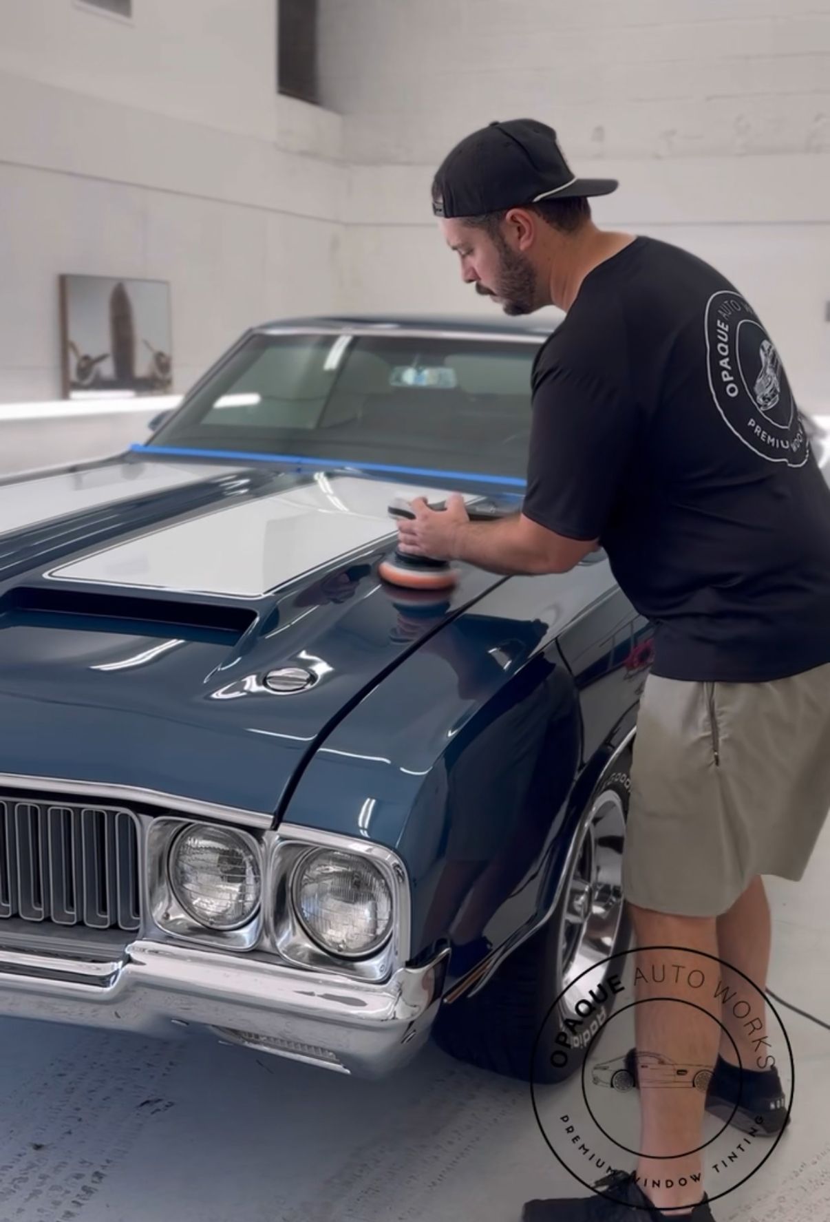 Man polishing a blue classic car hood in a well-lit garage; white racing stripes, black t-shirt, baseball cap.