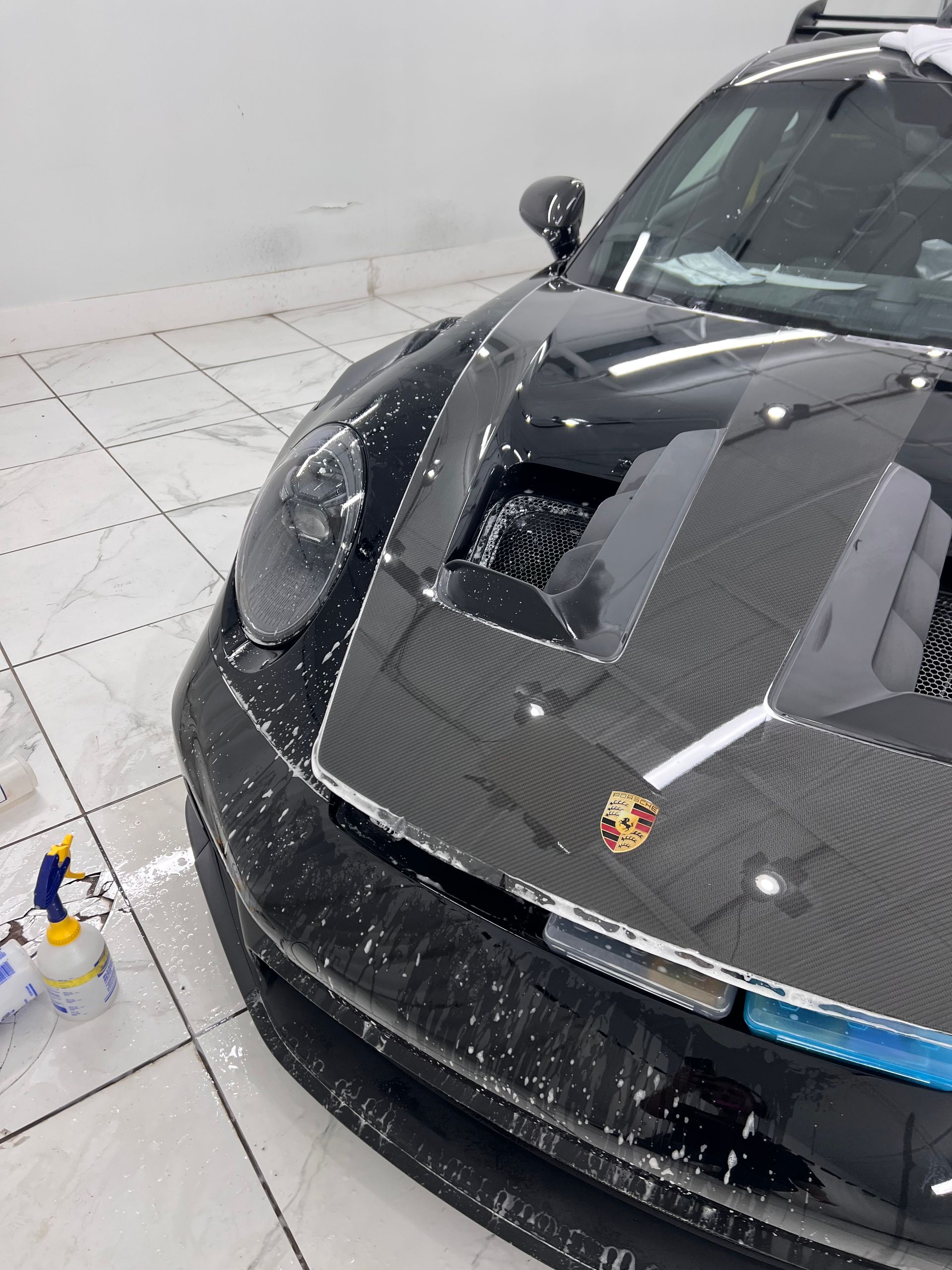 Black Porsche sports car with carbon fiber hood, wet from washing, in a tiled garage.
