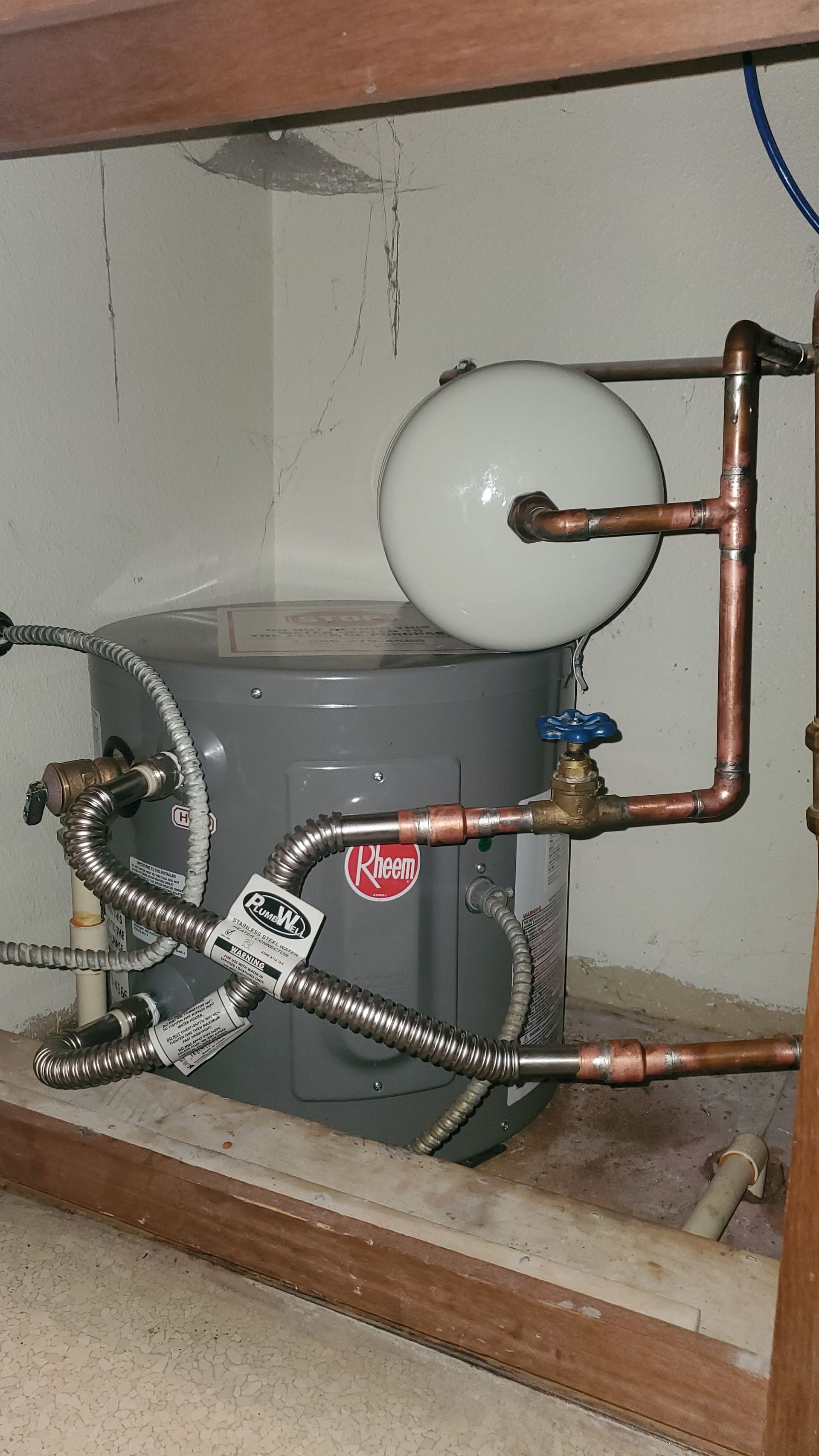 A Rheem water heater with copper pipes, a white expansion tank, and a blue valve in a wooden-framed enclosure.