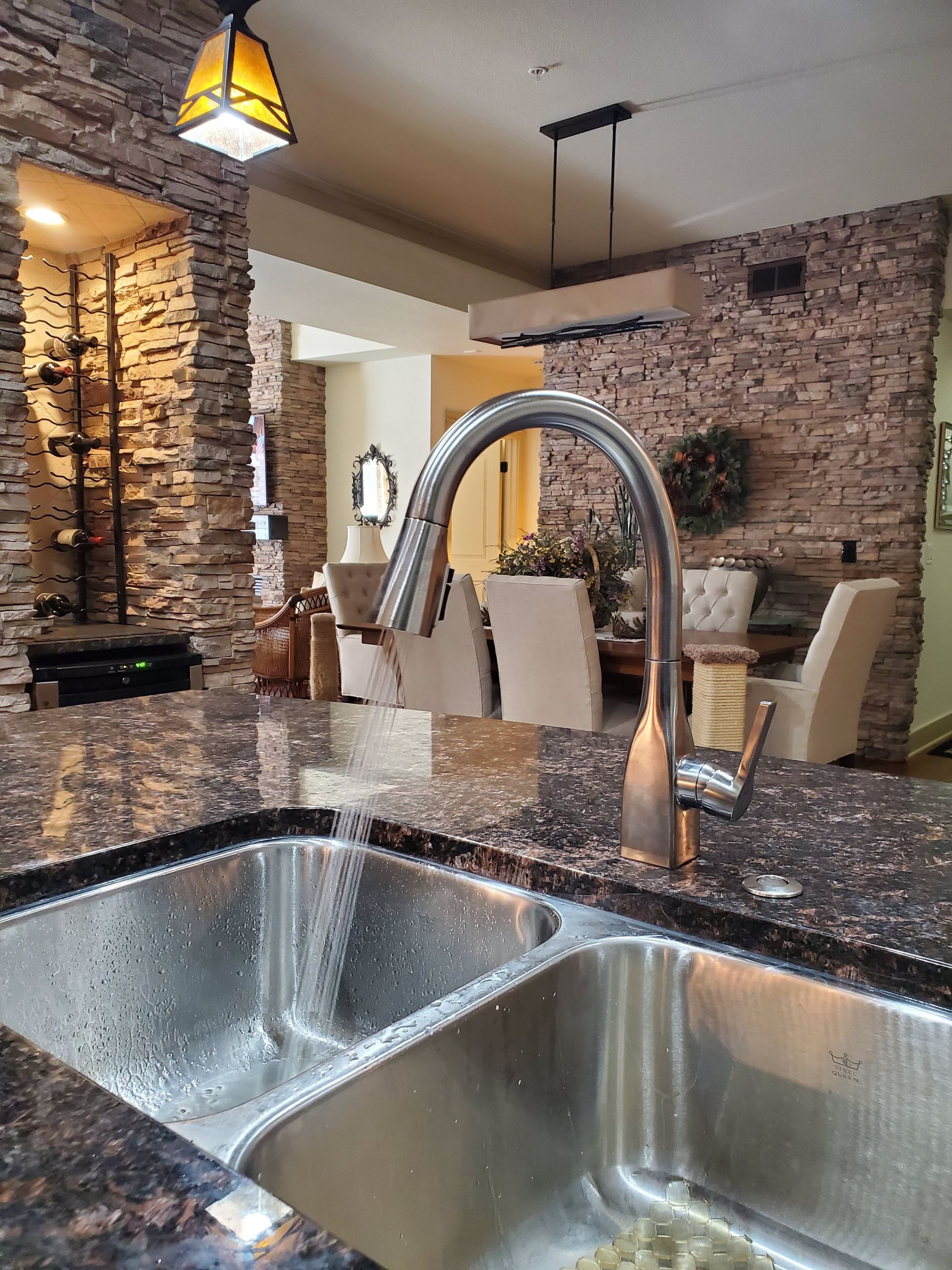 Stainless steel kitchen sink with faucet, granite countertop, stone wall background.