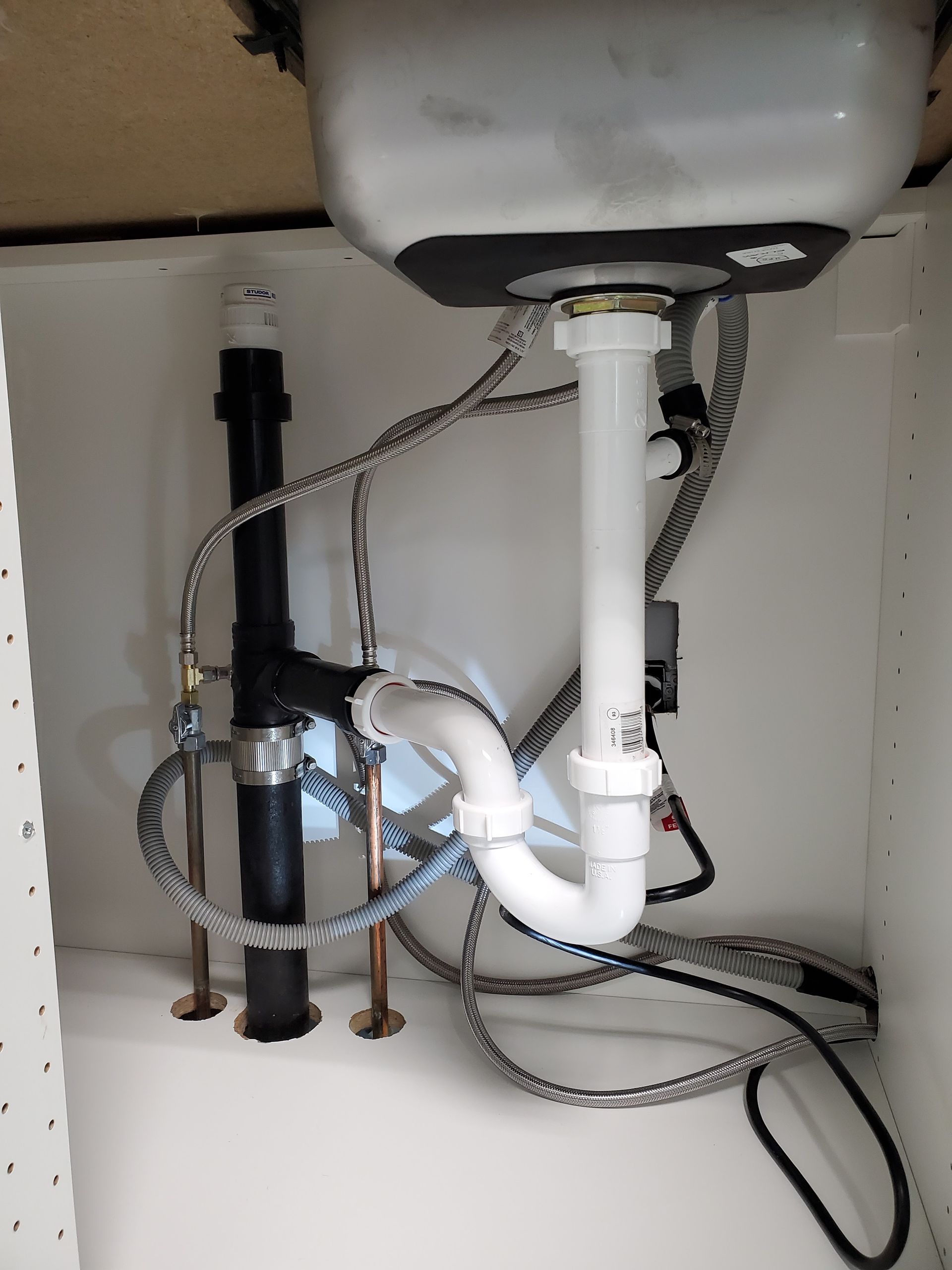 Plumbing under a white sink in a cabinet. White and black pipes, copper tubes, and water lines are visible.
