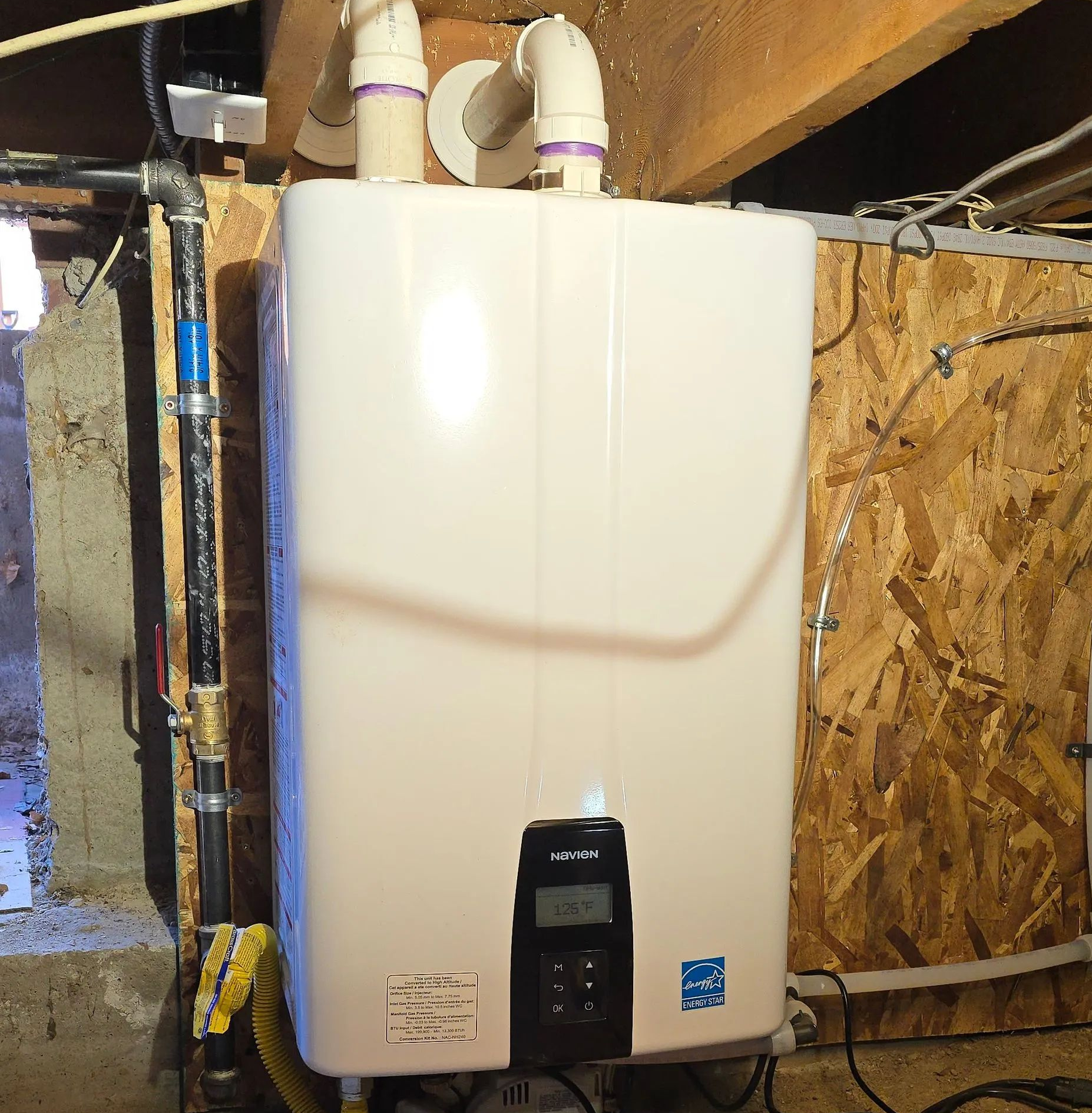 White wall-mounted water heater in a basement with visible pipes and plywood wall.
