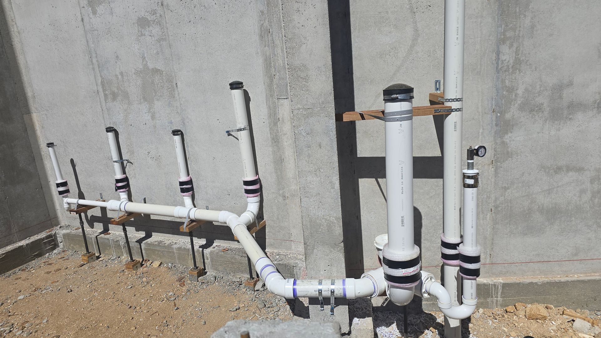 White PVC pipes against a concrete wall. Several pipes with black capped ends and a wooden brace are visible.