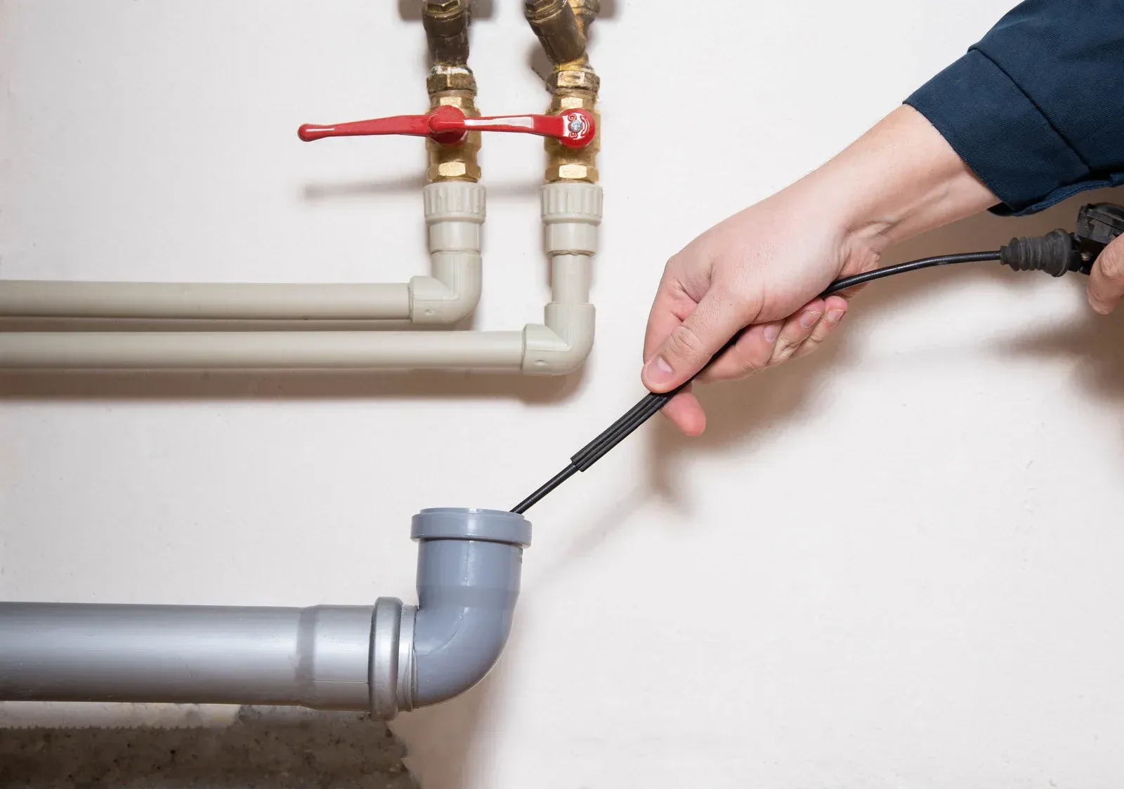 A hand feeds a flexible plumbing snake into a grey drain pipe beneath water valves.