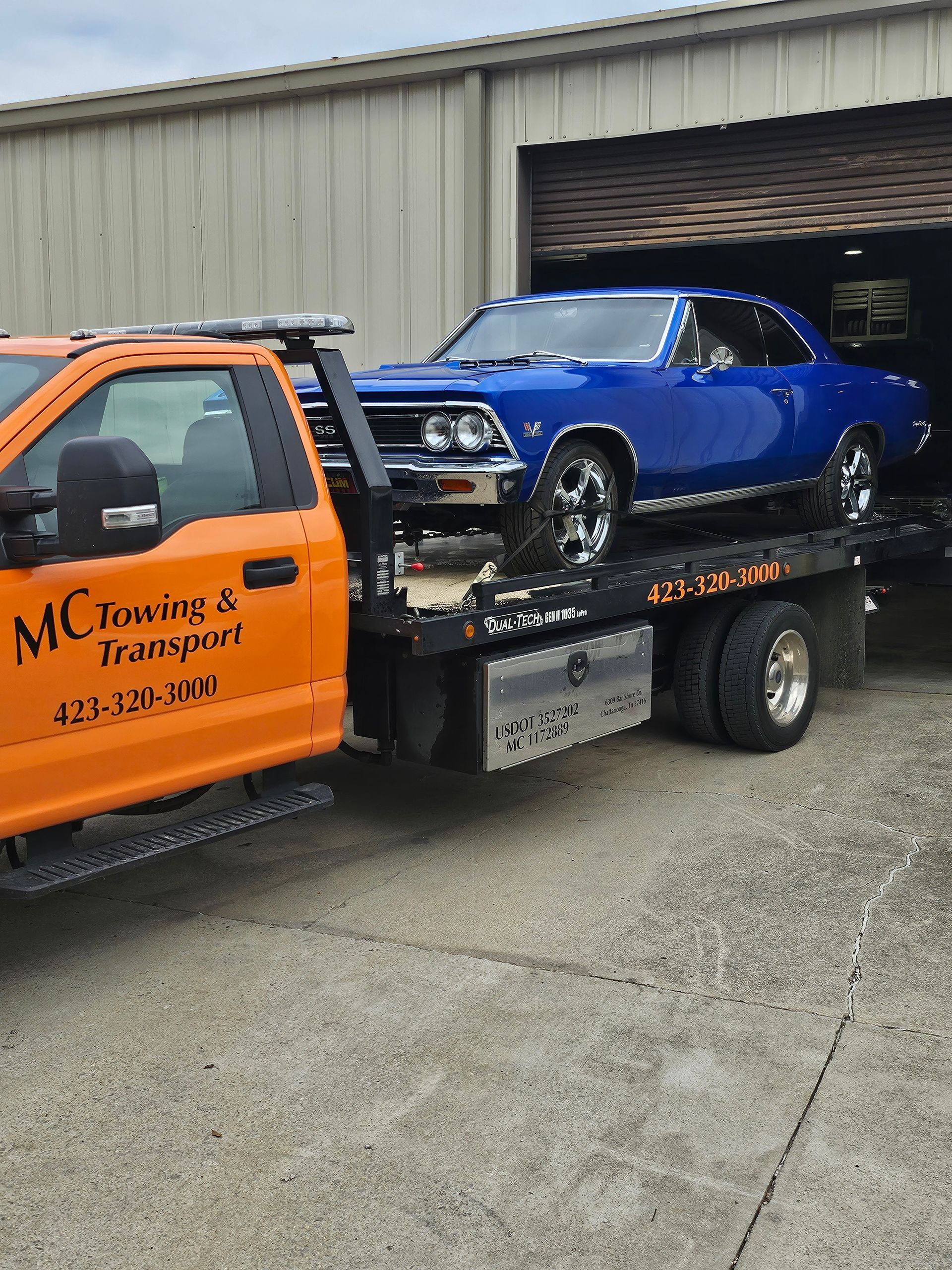 A bright blue classic muscle car parked on the back of an orange flatbed tow truck in front of a metal warehouse.