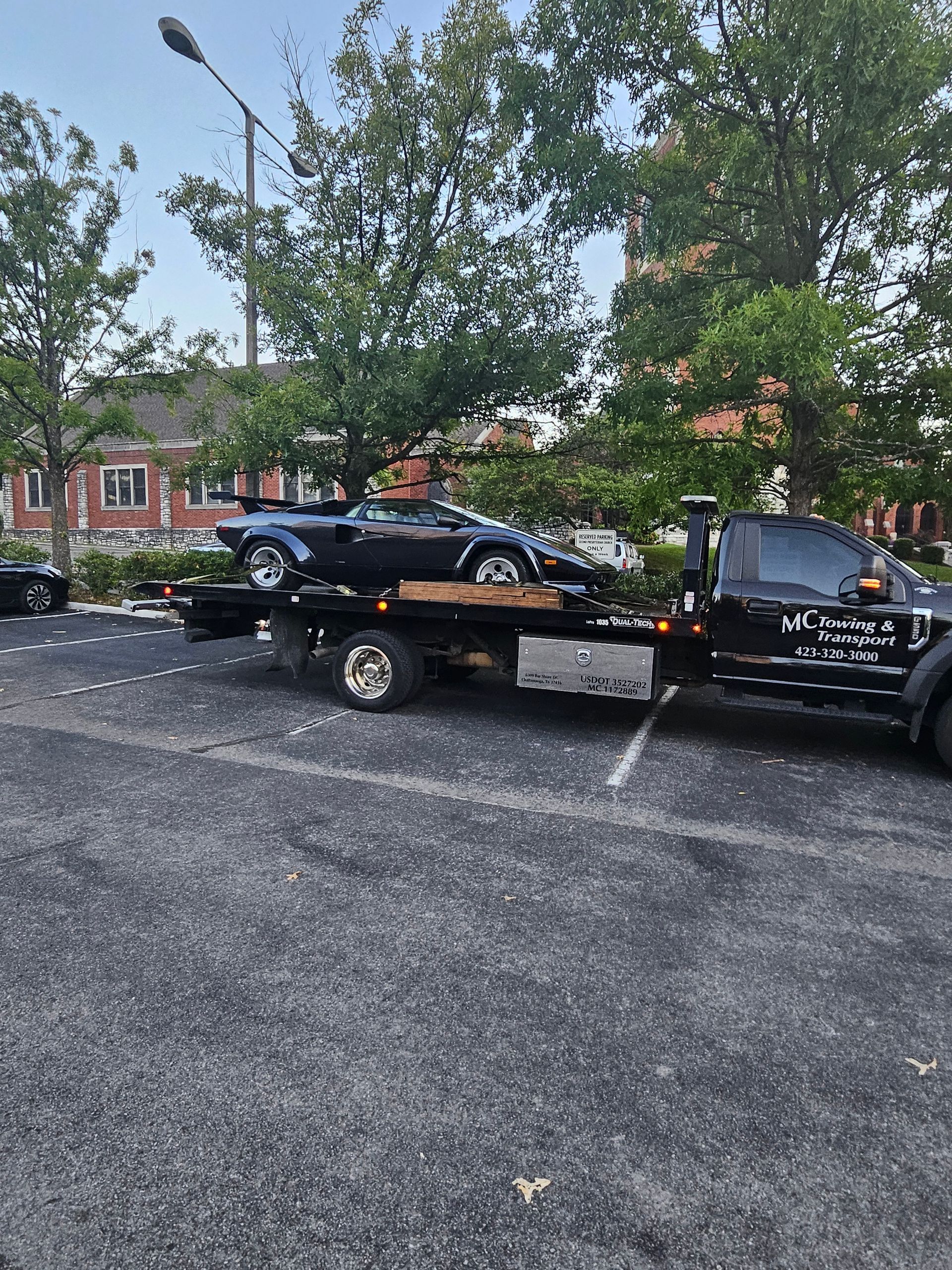A dark, wedge-shaped vintage sports car loaded on a flatbed tow truck in an outdoor parking lot with trees in the back.