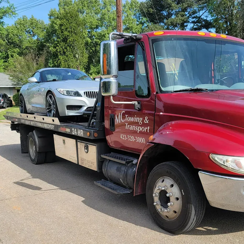 A red MC Towing & Transport flatbed tow truck carries a silver BMW convertible parked on a suburban street.