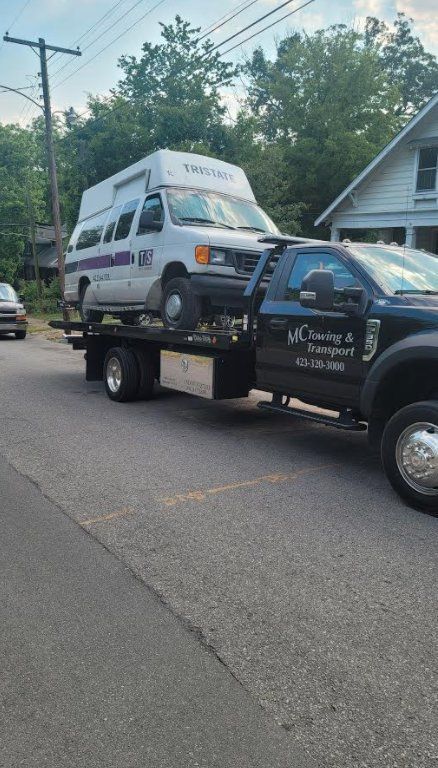 A white van with a high roof loaded onto a black flatbed tow truck on a suburban street.