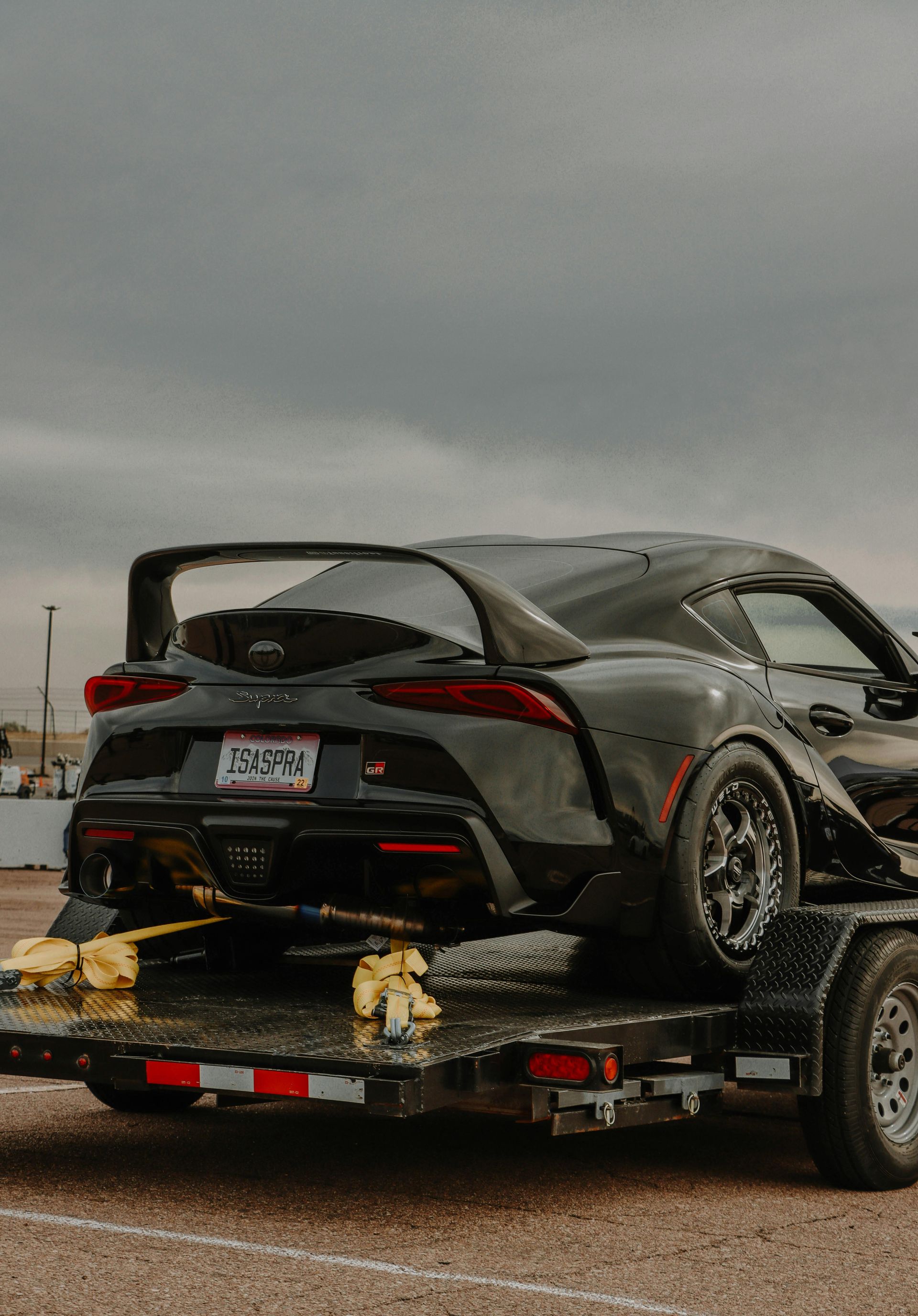 A dark gray Toyota Supra with custom racing wheels is securely strapped to a flatbed trailer outdoors on a cloudy day.