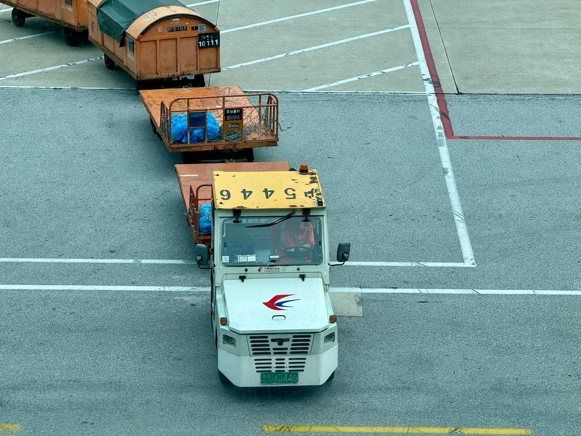 A white airport tug vehicle pulling orange luggage carts across an asphalt tarmac.
