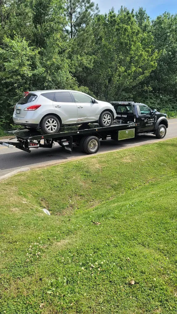 A silver crossover SUV is loaded on the flatbed of a black tow truck parked on the side of a road near a grassy ditch.