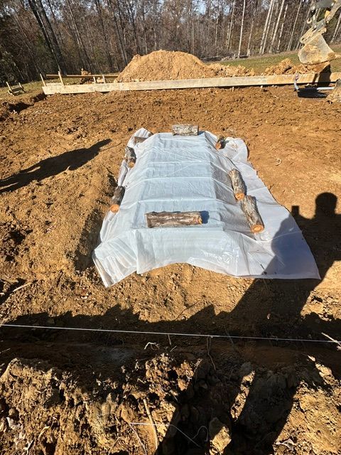 A tarp covers soil, held down by logs, in an outdoor setting with a pile of dirt in the background.