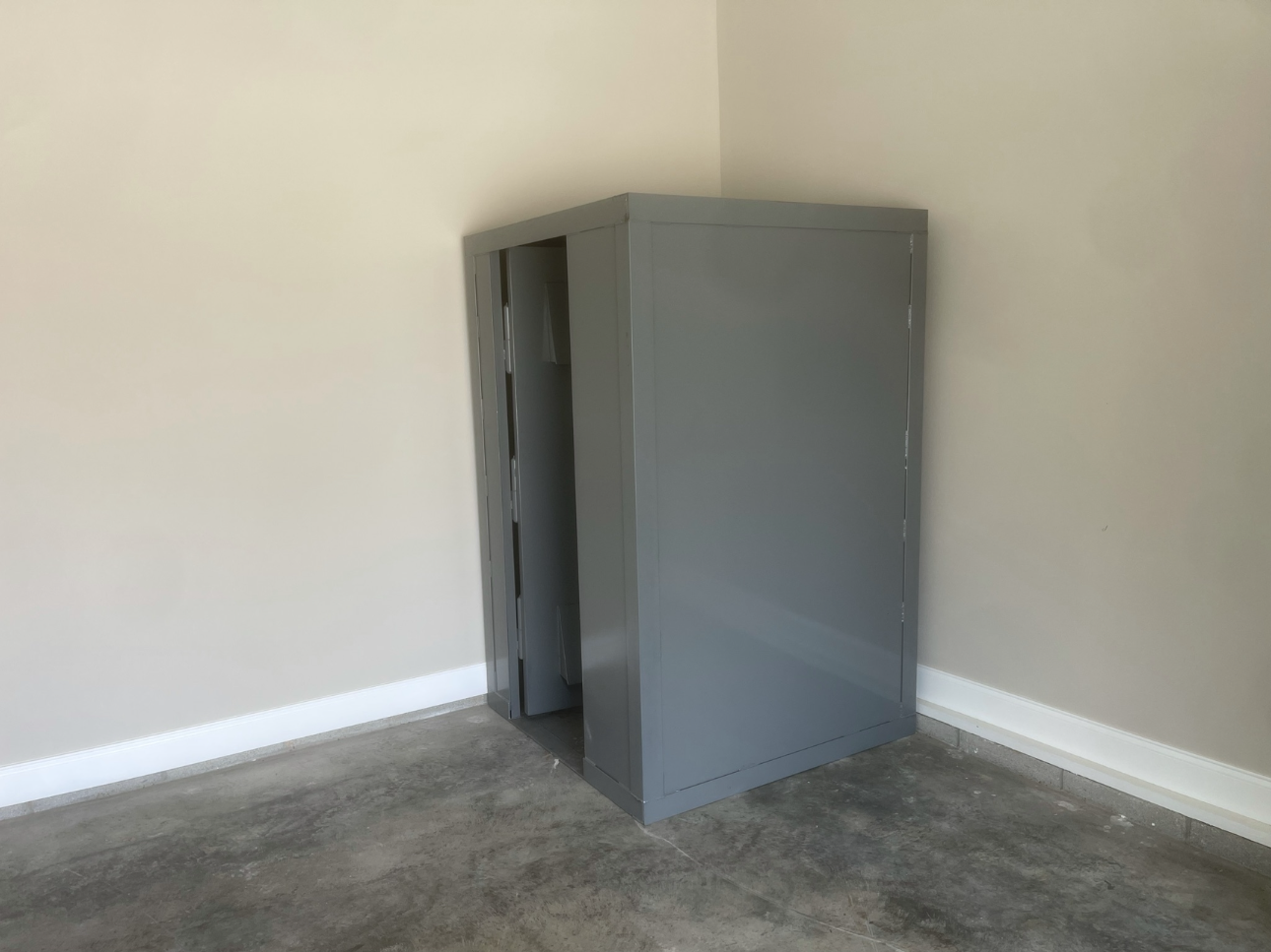 Gray metal cabinet in a corner of a room, walls painted beige, concrete floor, white baseboards.