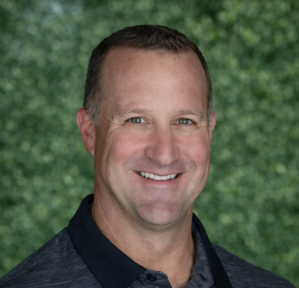 Headshot of a man with short brown hair smiling, wearing a dark gray polo shirt against a blurry green background.