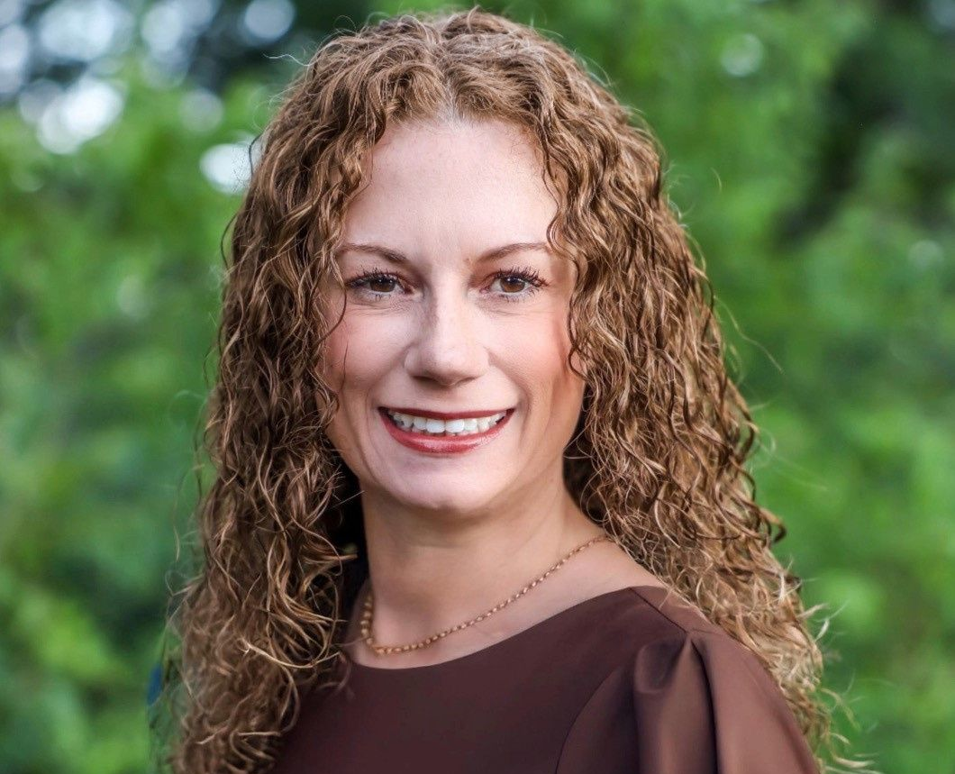 A professional headshot of a person with curly auburn hair smiling, wearing a brown top against a blurred green background.