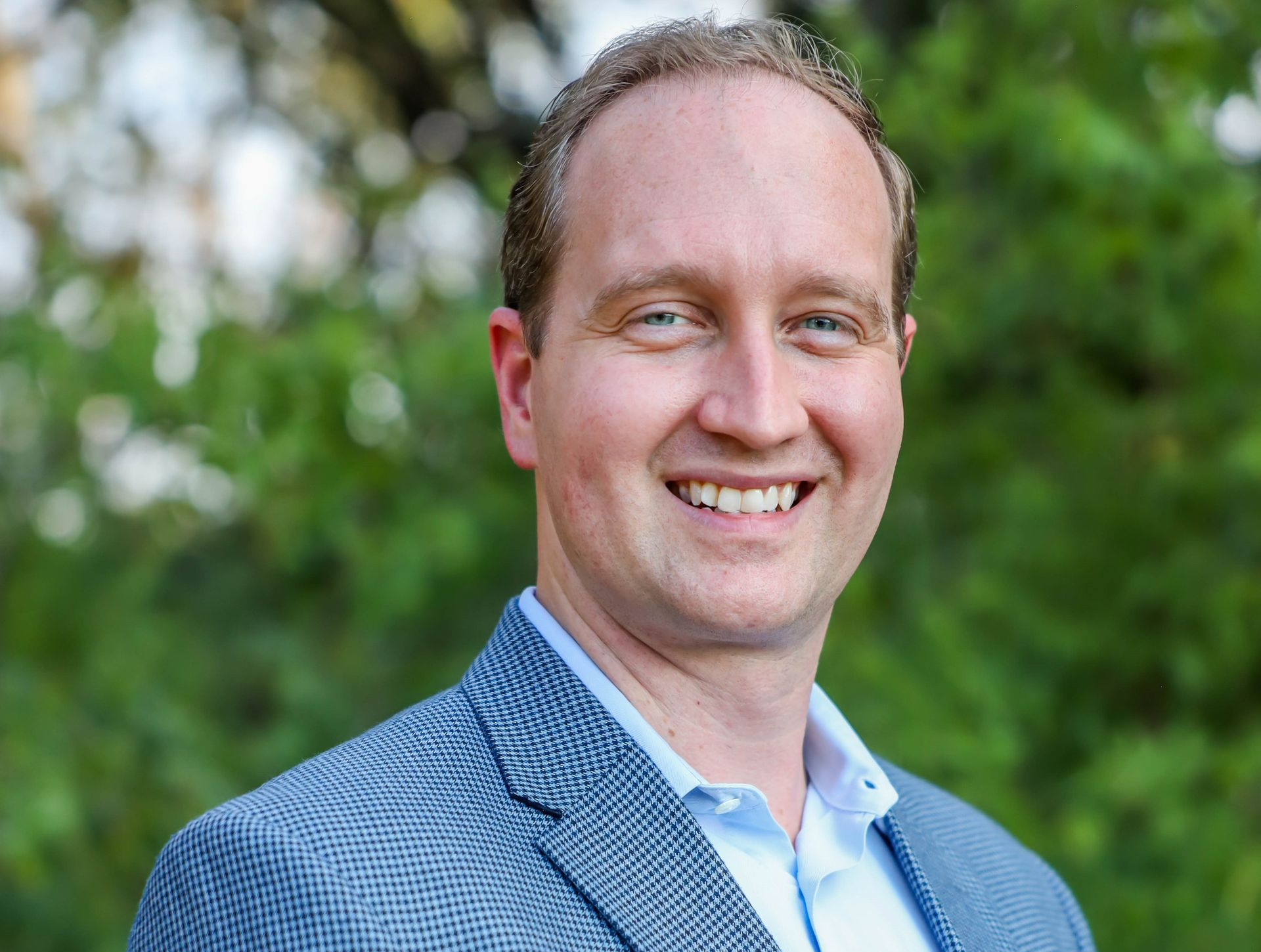 A smiling person in a blue patterned blazer and light blue button-down shirt, set against a blurry green foliage background.