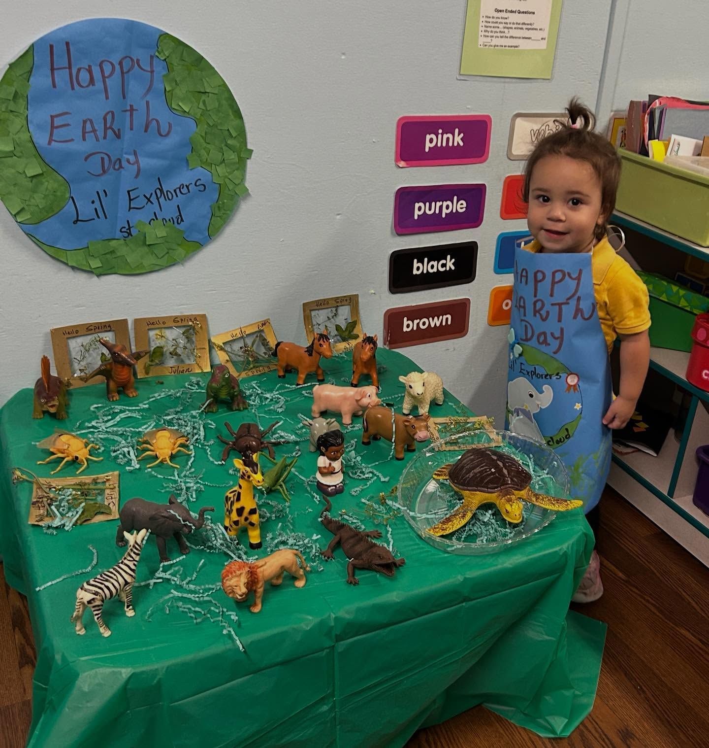 Child beside Earth Day display with green table, animal figures, and “Happy Earth Day” sign
