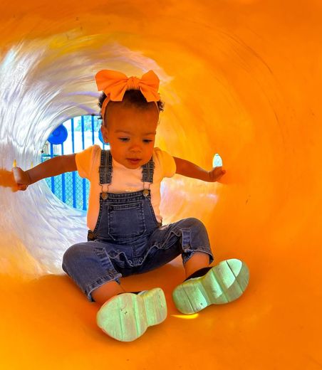 Toddler in denim overalls and orange bow sliding through a bright orange tunnel