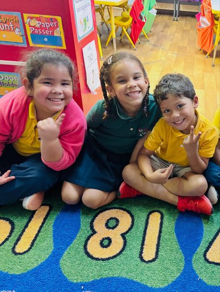 Three children smiling on a colorful classroom rug with number blocks nearby