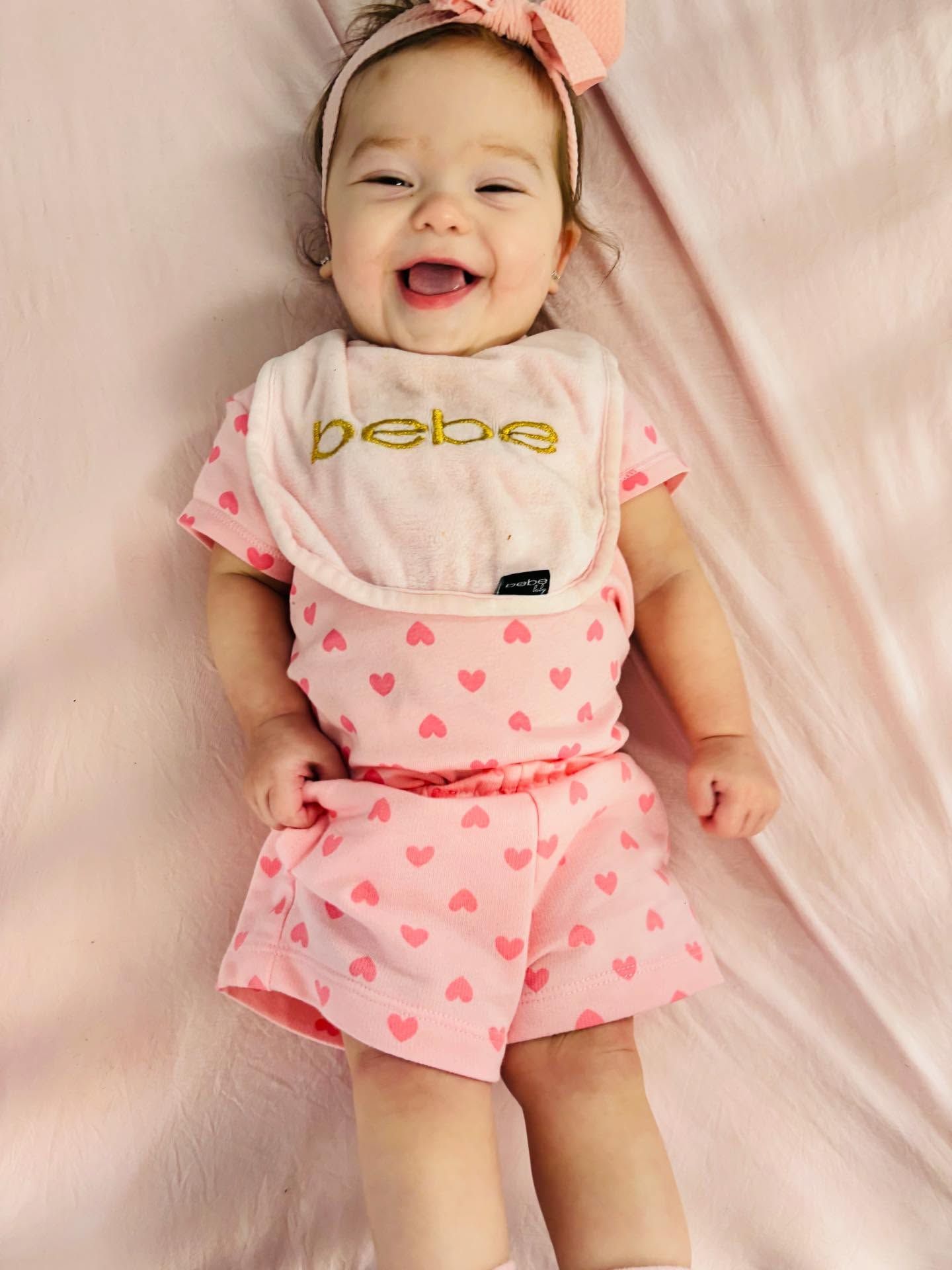 Smiling baby lying on a pink bedspread, wearing a pink heart outfit and bib.
