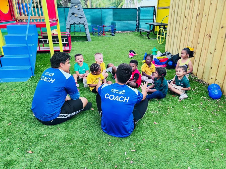 Two coaches sit on grass, leading a group of children in a colorful outdoor play area.