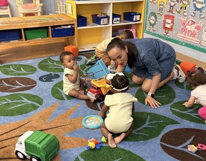 Teacher kneels with toddlers on a colorful classroom rug, playing with toys together.