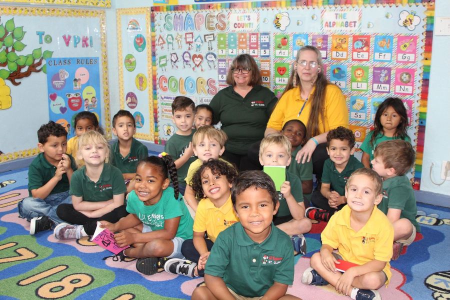 Classroom group photo with children seated on a colorful rug and two adults standing behind them