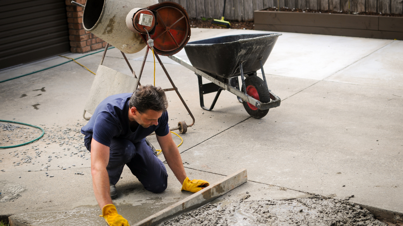 Man leveling concrete with a tool, next to a cement mixer and wheelbarrow on a driveway.