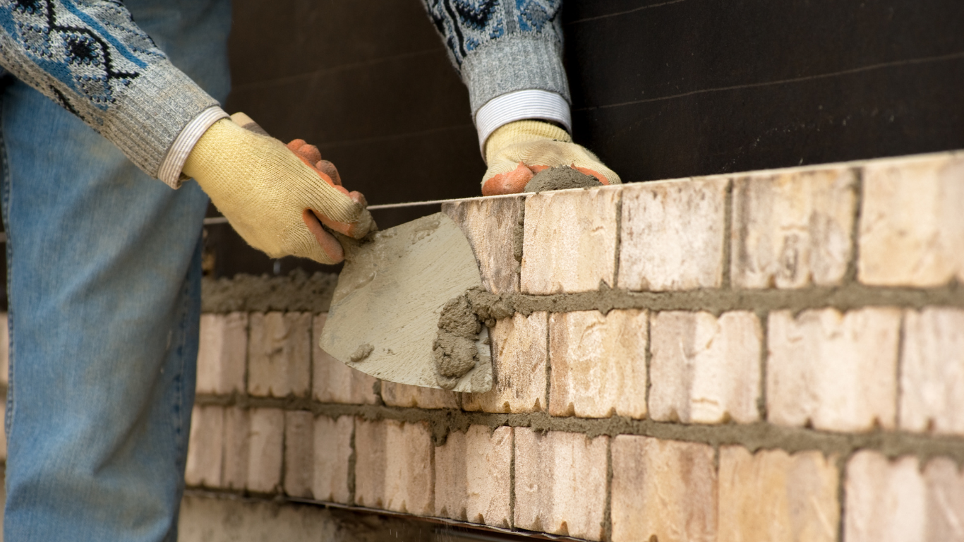 Person in jeans and sweater using a trowel to apply mortar between bricks, building a wall.