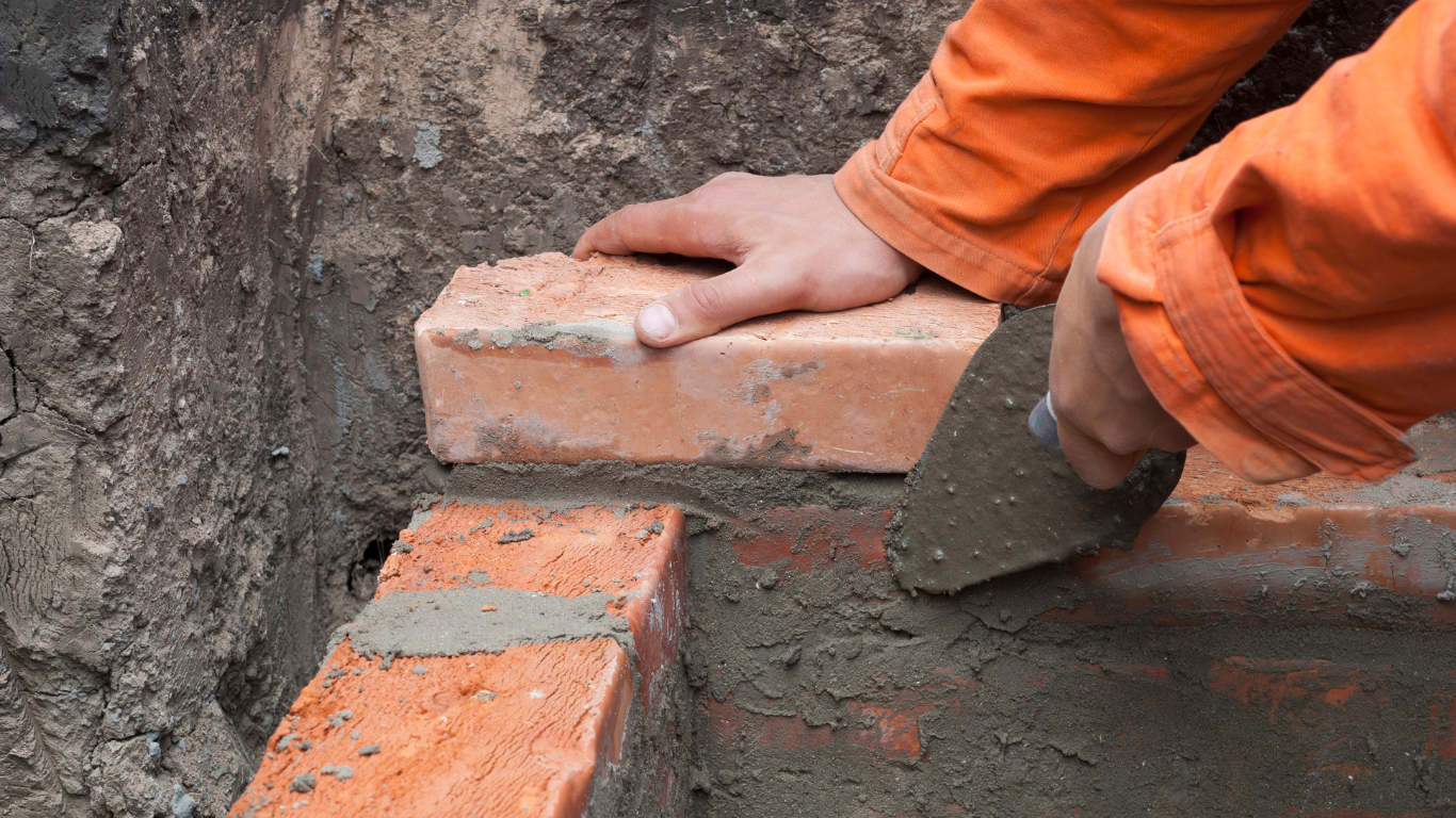 A person laying a brick, using a trowel to spread mortar in a dirt setting.