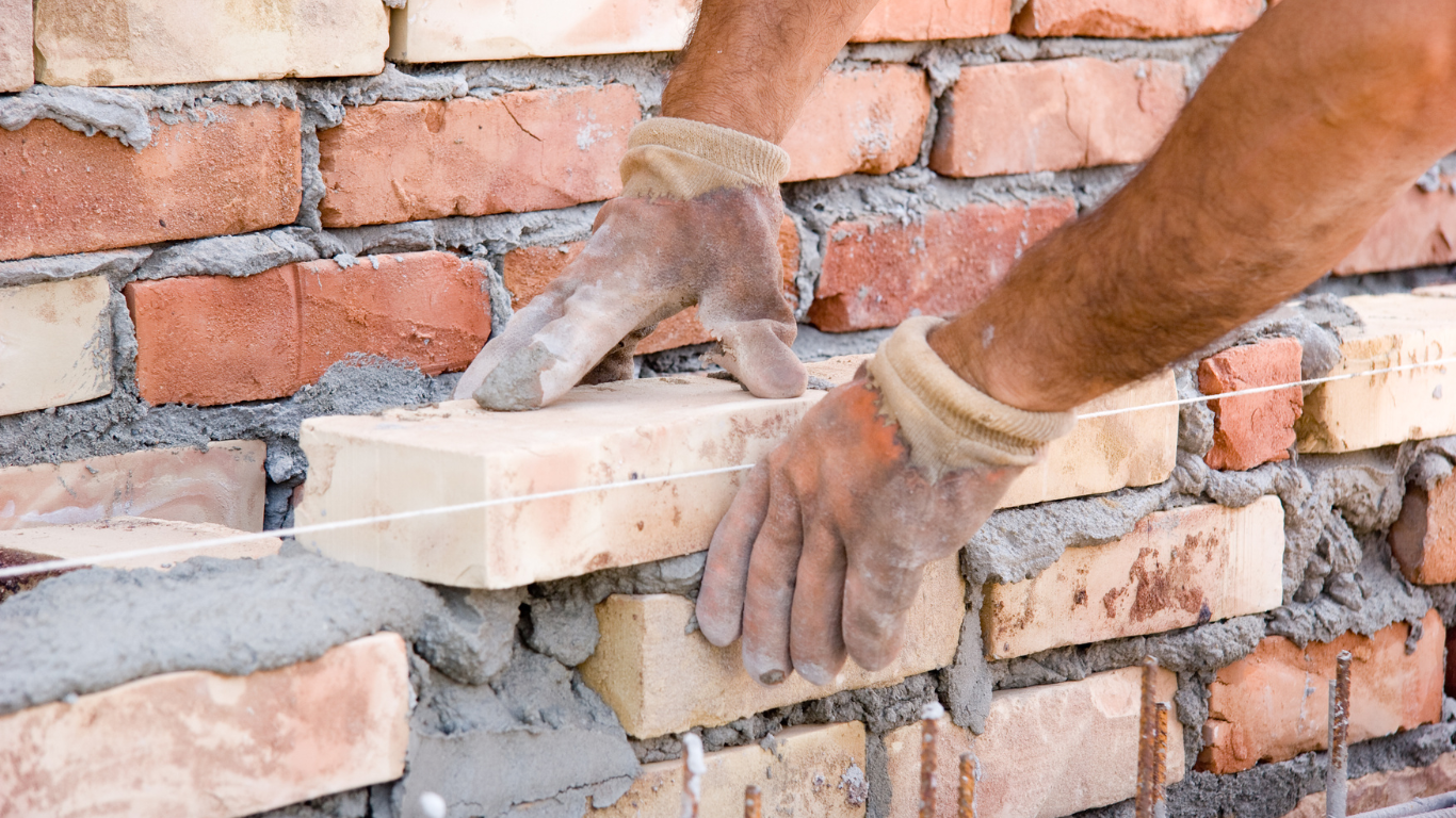 Person laying a brick, hands covered in mortar, on a partially built brick wall.