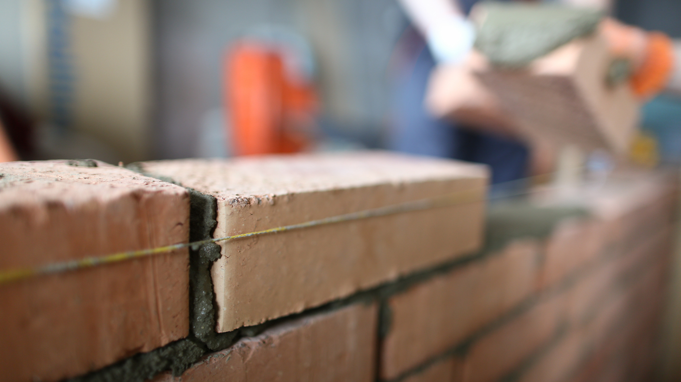 Close-up of bricklaying with mortar and a level string; an orange bucket is in the background.