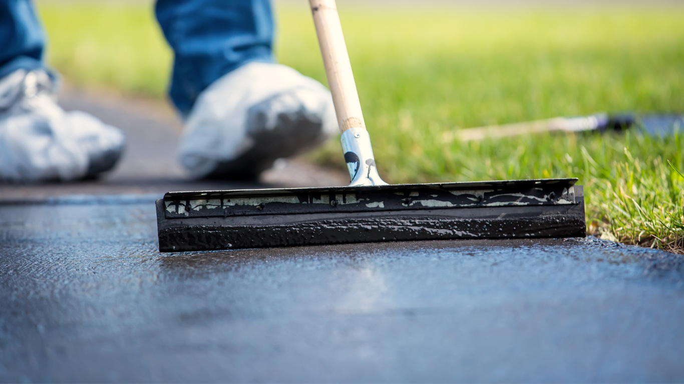 Person using a squeegee to apply black sealant to a paved surface outdoors.