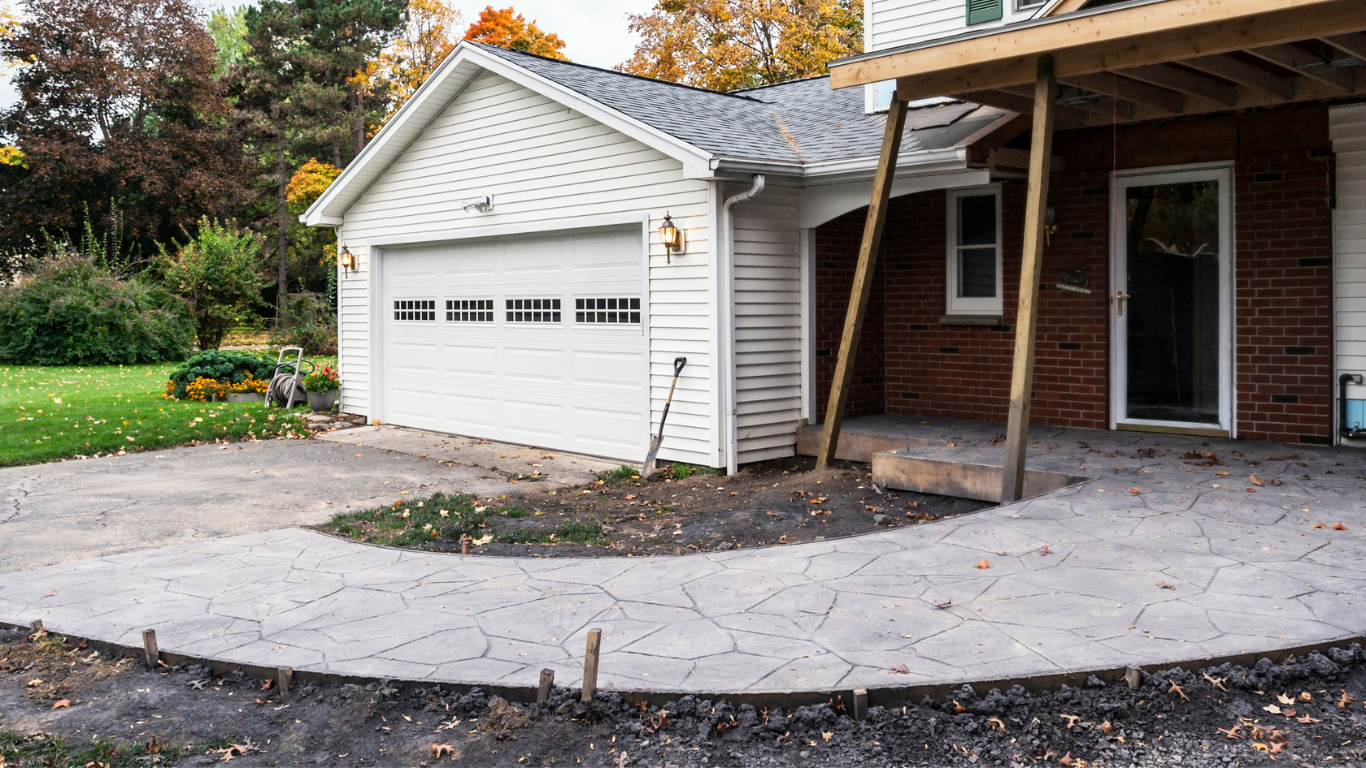 A driveway curves past a white garage and red brick house with a covered porch; fall foliage surrounds.