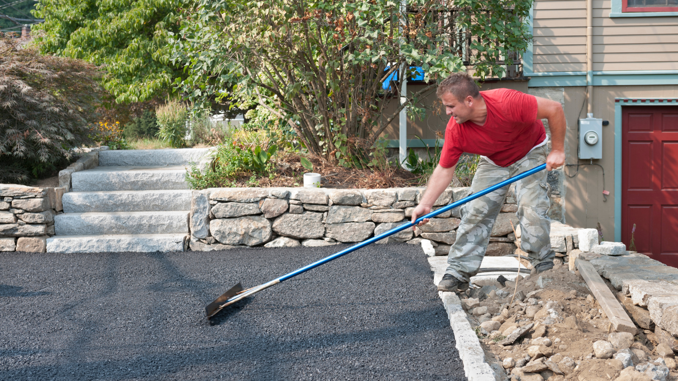 Man in red shirt rakes asphalt driveway next to a retaining wall and house.
