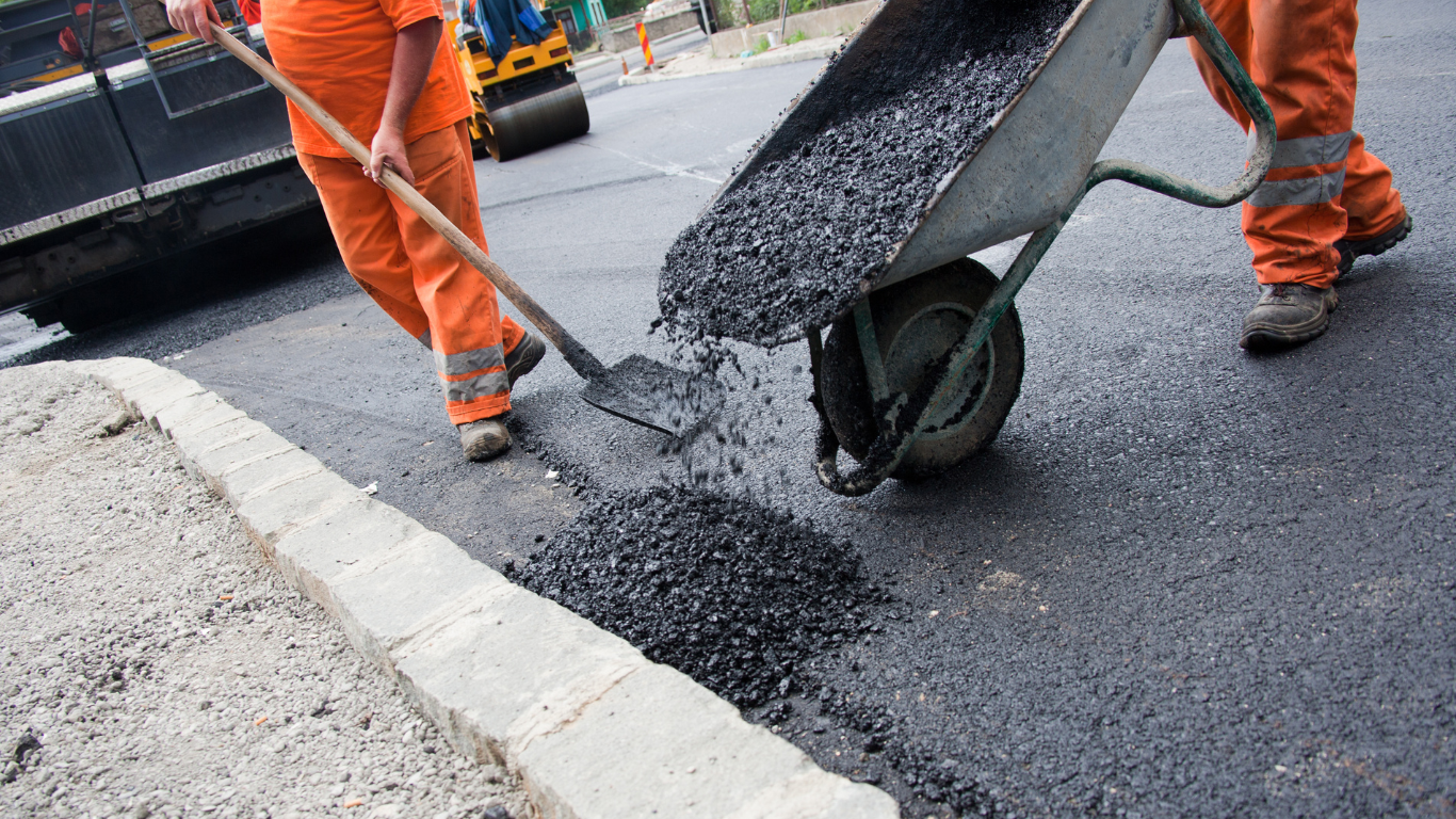 Road workers in orange coveralls paving a road with asphalt.
