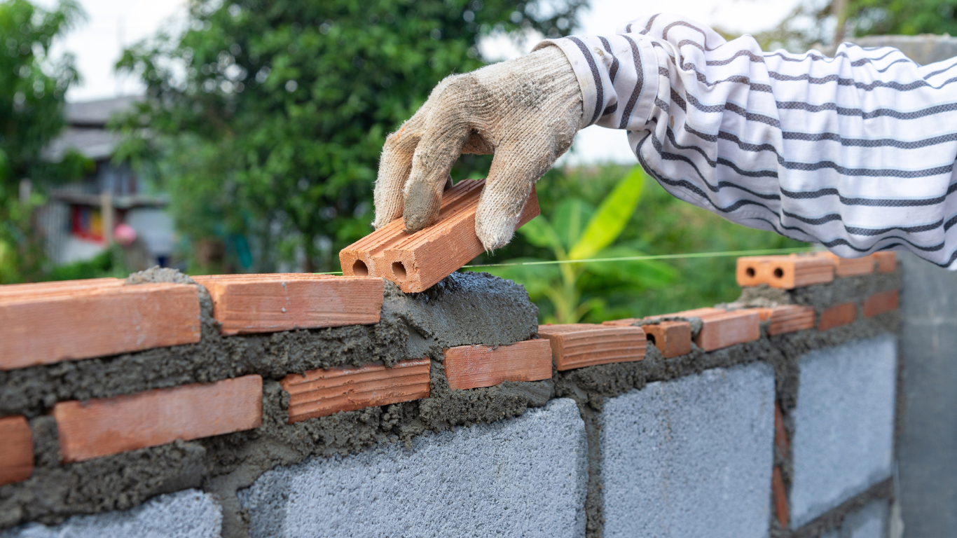 Person wearing gloves laying a red brick onto mortar on top of a gray concrete block wall.